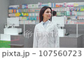 Female pharmacist posing with crossed arms. Woman druggist standing at workplace. Smiling woman wearing robe and standing at background of medicine and colleague. Pharmacists in pharmacy. 107560723