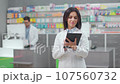 Female pharmacist holding tablet at foreground. Woman druggist working with gadget in pharmacy. Woman in white robe standing at background of showcase and colleague. Pharmacists in drugstore. 107560732