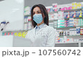 Female pharmacist looking at computer. Woman pharmacist working with cash register. Druggist wearing white robe and protective mask. Showcase with pills and syrups. Caucasian pharmacist in pharmacy. 107560742