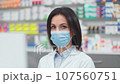Close up view of female druggist looking at camera. Portrait of woman pharmacist in pharmacy. Woman wearing robe and protective mask. Showcase with pills and syrups. Caucasian pharmacist in drugstore. 107560751