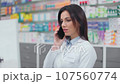 Female pharmacist looking at computer monitor. Woman pharmacist communicating with client by phone. Woman in white robe talking by phone. Showcase with medicine. Caucasian druggist in drugstore. 107560774
