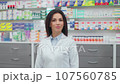 Smiling female druggist with crossed arms. Woman pharmacist at workplace. Woman in white robe standing at background of showcase with medicine. Cheeful pharmacist in drugstore. 107560785