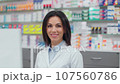 Smiling female pharmacist looking at camera. Woman pharmacist at workplace. Woman in white robe standing at background of showcase with medicine. Cheeful pharmacist in drugstore. 107560786