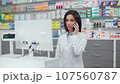 Female pharmacist looking at computer monitor. Woman pharmacist working with cash register. Woman in white robe talking by phone. Showcase with medicine. Caucasian pharmacist in pharmacy. 107560787