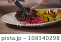 A man cuts meat in a plate and eats fried potatoes with grilled vegetables in a restaurant, close-up. 108340298