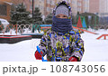 3 years old boy enjoying winter day on a fenced sports ground. He approaches the camera with a baby shovel in his hand wrapped in a warm scarf. Fun winter holidays. Holiday outdoors in cold winter 108743056