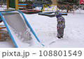 3 years old boy enjoying winter day on a fenced sports ground. He approaches the camera wrapped in a warm scarf. Fun winter holidays. Holiday outdoors in cold winter 108801549