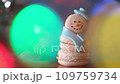 Snowman dessert macaron decorated with hat and scarf with colorful lights and flashing bokeh on foreground and Christmas tree on background. Merry Christmas and Happy New Year festive concept 109759734