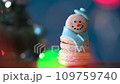 Snowman dessert macaron decorated with hat and scarf with colorful lights and flashing bokeh on foreground and Christmas tree on background. Merry Christmas and Happy New Year festive concept 109759740