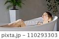 Stunning woman resting comfortably in white glossy tub, gently washing skin. Side view of enjoyed bare girl taking bath in stylish, brightly lit bathroom, in motion. Pleasure time, self care concept. 110293303