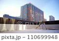 Apartment with an outdoor terrace. Bearded man in a warm jacket cleans snow with snow shovel outdoors in a private area. Snow removal in sunny winter day. A high-rise building in the background. 110699948