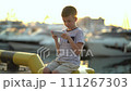 An eight-year-old boy sits on the shore and plays the phone against the backdrop of yachts and boats. 111267303