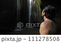 A young topless man with abundant hair on his shoulders and back stands alone at the waterfall and admires its view, looking up from the bottom of the river canyon. Rear view 111278508