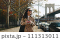 Stylish female texting via smartphone and smiling, while walking with the iconic Brooklyn Bridge in the back 111313011