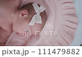 Portrait of a beautiful newborn baby girl lying in a white cot under a pink plaid with ruffles at home with a bow on her head and looking around. 111479882