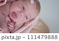 Portrait of a beautiful newborn baby girl lying in a white cot under a pink plaid with ruffles at home with a bow on her head and looking around. Vertical 111479888
