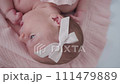 Portrait of a beautiful newborn baby girl lying in a white cot under a pink plaid with ruffles at home with a bow on her head and looking around. 111479889
