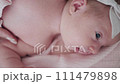 Portrait of a beautiful newborn baby girl lying in a white cot under a pink plaid with ruffles at home with a bow on her head and looking around. 111479898