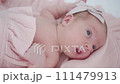 Portrait of a beautiful newborn baby girl lying in a white cot under a pink plaid with ruffles at home with a bow on her head and looking around. 111479913