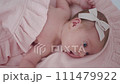 Portrait of a beautiful newborn baby girl lying in a white cot under a pink plaid with ruffles at home with a bow on her head and looking around. 111479922