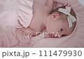 Portrait of a beautiful newborn baby girl lying in a white cot under a pink plaid with ruffles at home with a bow on her head and looking around. 111479930