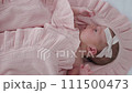 Portrait of a beautiful newborn baby girl lying in a white cot under a pink plaid with ruffles at home with a bow on her head and looking around. 111500473