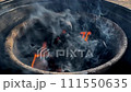 Vivid orange flames lick through the dense smoke over the hot coals of a charcoal grill. 111550635