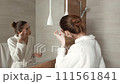 A woman in a white robe is washing her face, standing before a large mirror in a well-lit, contemporary bathroom 111561841