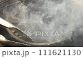 Metal tongs move a bowl of charcoal grill, covered in grey ash and exuding thick smoke. 111621130