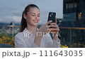Woman smiling with phone in hand chatting light from cell phone, outside glowing buildings at night against big city skyscraper background, communication and internet while traveling. 111643035