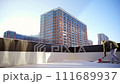 Apartment with an outdoor terrace. Bearded man in a warm jacket cleans snow with snow shovel outdoors in a private area. Snow removal in sunny winter day. A high-rise building in the background. 111689937