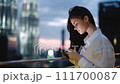 A woman looks at her phone, smiling and laughing typing a message backdrop of a nighttime city and glowing buildings on the roof of a skyscraper, hair flying in the breeze, work and leisure city life. 111700087