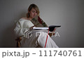medium-sized woman in light clothes reads a book while sitting in a white chair in a light room 111740761
