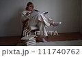 medium-sized woman in light clothes reads a book while sitting in a white chair in a light room 111740762