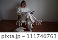 medium-sized woman in light clothes reads a book while sitting in a white chair in a light room 111740763