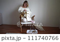 mid size woman in light clothes embroiders a picture in gold and beige tones while sitting on a white chair in a light room 111740766