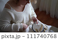 mid size woman in light clothes embroiders a picture in gold and beige tones while sitting on a white chair in a light room 111740768