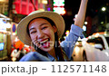 Smiling Asian woman in straw hat greeting with a wave, city nightlife sparkling in the background. Female traveler backpacker in Bangkok, Thailand. 112571148