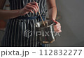A woman in a striped apron makes wax candles. She stirs the melted wax in a pan. The creative process of making wax scented candles. 112832277
