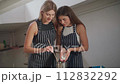 Two young attractive women in striped aprons are making scented wax candles. One of them adds the wax to the pan, the other stirs it. The process of making aromatic wax candles. 112832292