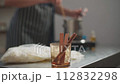 A woman in a striped apron makes wax candles. She stirs the melted wax in a pan and monitors the temperature of the wax with a thermometer. 112832298