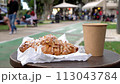 Coffee in a paper cup and a bun on the table of a street cafe with people walking and riding scooters on the boulevard on the background 113043784