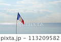 A small French flag on a thin flagpole flutters over the sea 113209582