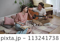 A guy and a girl are sitting on a plaid on a wooden floor in the living room. The guy looks at the girl and plays the acoustic guitar, the girl flips through the book and looks at the guy with a smile 113241738