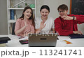 Students communicate using a laptop and the Internet. A guy and two girls look into the laptop camera, smile, wave their hands and read notes from a notepad, sitting at a table in the living room. 113241741