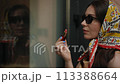 Slow motion. Close-up. Stylish woman in a headscarf and sunglasses looks at her reflection in a store window and paints her lips with lipstick while standing outdoors on a summer day. 113388664