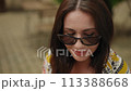 Slow motion. Close-up. A woman in dark-framed sunglasses poses for the camera, putting a headscarf on her head. A woman looks at the camera over her glasses and smiles while outdoors on a summer day. 113388668