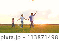 Mother father and daughter playing flying kite at sunset natural field green grass. Happy family parents and girl kid child holding hands relaxing together outdoor meadow flight toy entertainment 113871498