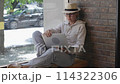 Wearing hat and eyeglasses woman sits on windowsill in cafe opens laptop starts working. Woman opens laptop to start online work taking advantage of cozy ambiance cafe Online modern work. 114322306