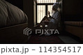 Woman in dark room sitting in robe by window. Attractive young girl in bathrobe and with towel on her head is holding cup, looking at window and smiling while sitting on window sill in old hotel room 114354517